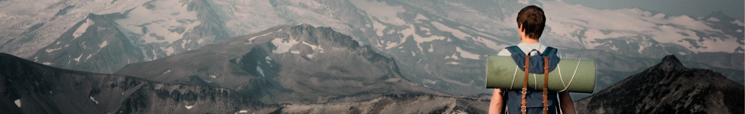 Person with backpack facing snow-capped mountains from a rocky peak